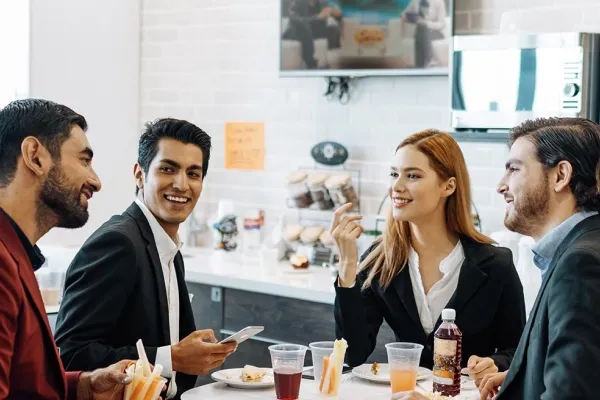 Four diverse business professionals—two men in suits, one man in a blazer, and one woman—sit at a table in a bright cafeteria or break room, talking and smiling during a lunch break.